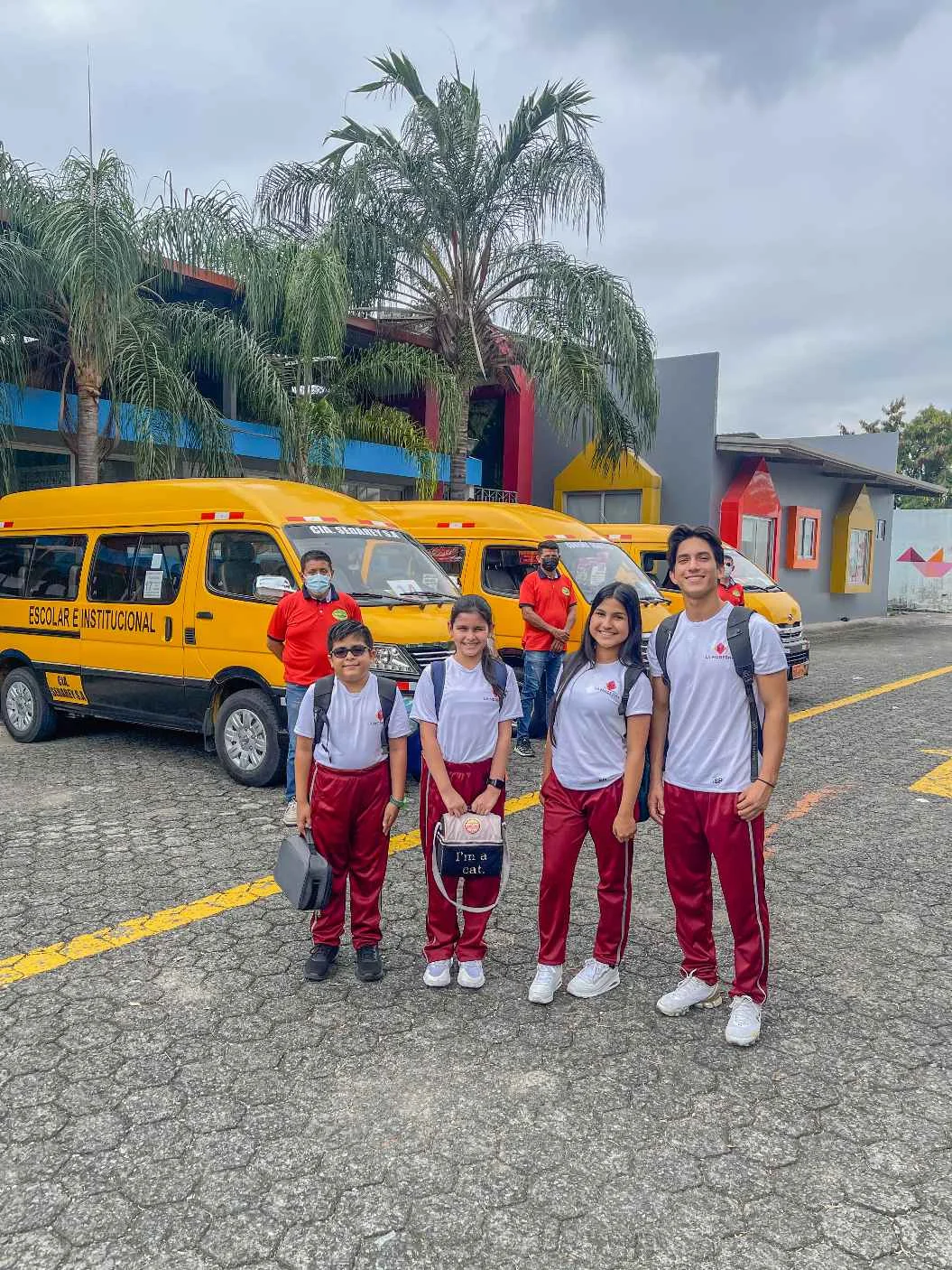 Four students in white and maroon uniforms smiling in front of yellow school buses at La Moderna campus pickup area