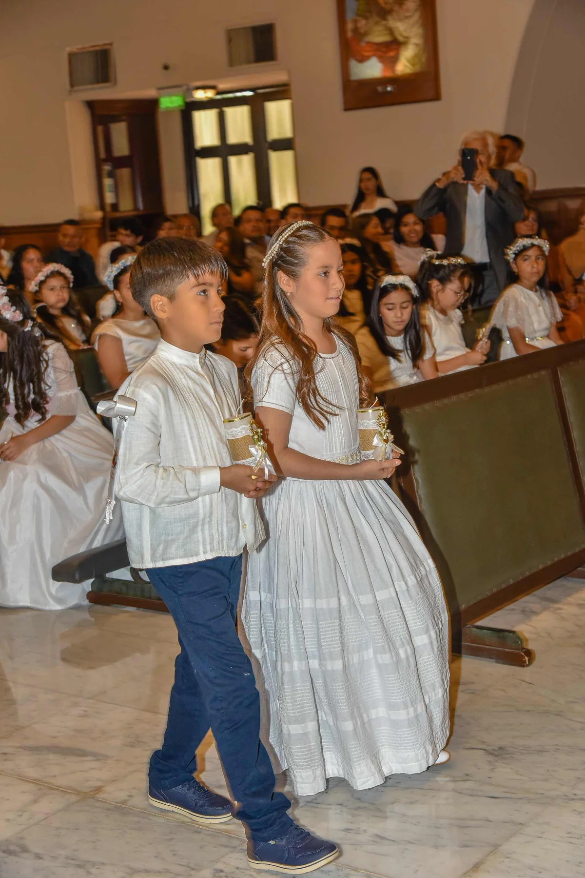 Two children in formal white attire walk down a church aisle holding decorated candles during a First Communion ceremony
