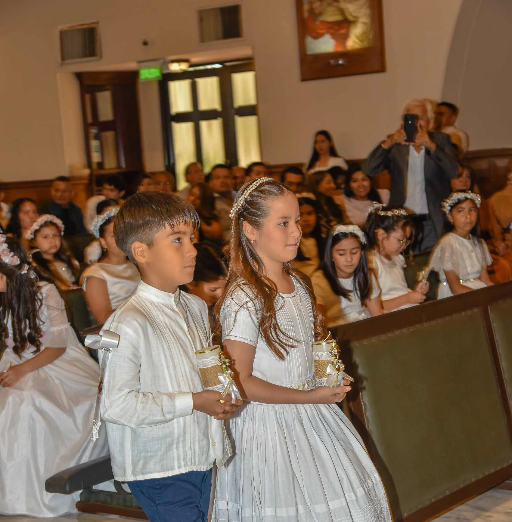 Two children in formal white attire walk down a church aisle holding decorated candles during a First Communion ceremony